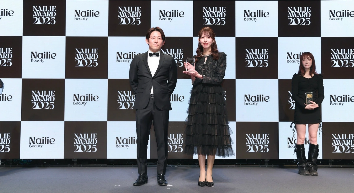 「Nailie AWARD 2025」のロゴが入った背景の前で、男女3名がステージに立っています。中央の男女はトロフィーを持っており、授賞式か表彰イベントの様子です。