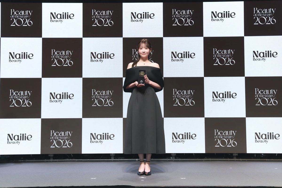 黒いオフショルダーのドレスを着た若い女性がステージ上でトロフィーを手にしています。背景には「Beauty of the year 2026」と「Nailie Beauty」のロゴがチェック柄に配置されており、美容関連の授賞式での一幕を捉えたものです。