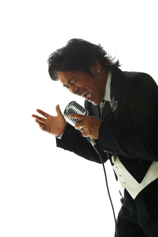 黒いタキシードと白いベストを着た男性が、ヴィンテージマイクに向かって情熱的に歌っている様子