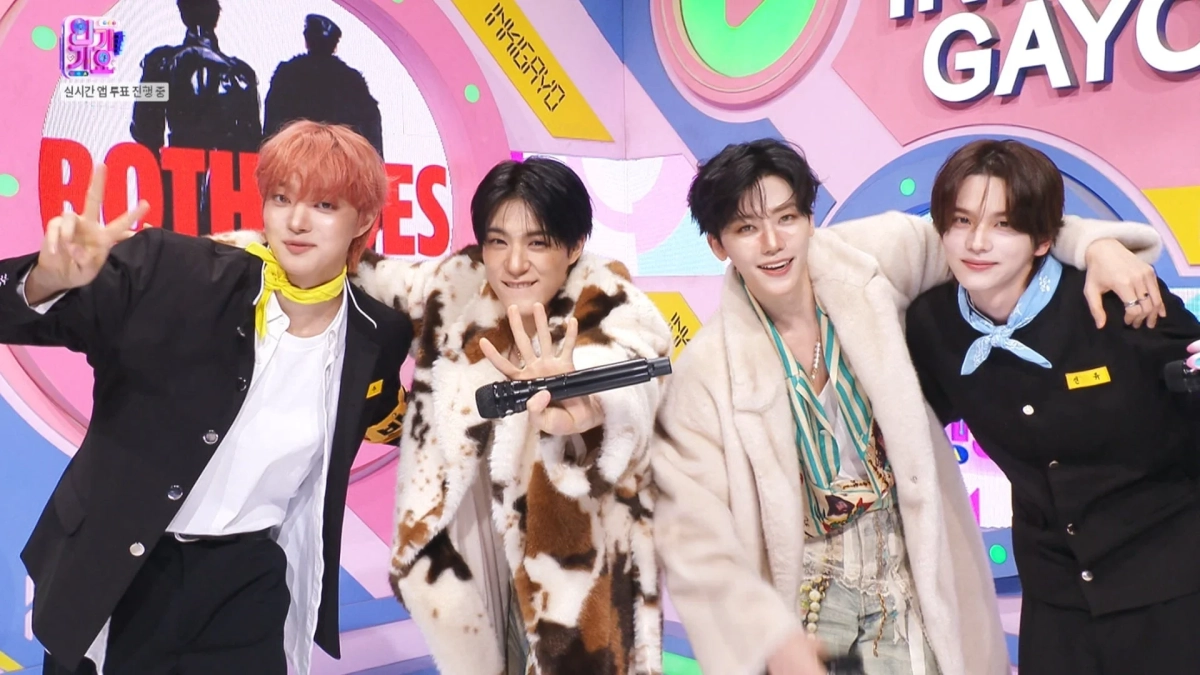 K-POPアイドルグループのメンバー4人が、カラフルなステージでポーズをとっています。個性的な衣装を身につけ、笑顔でカメラを見つめており、マイクを持ったメンバーもいます。