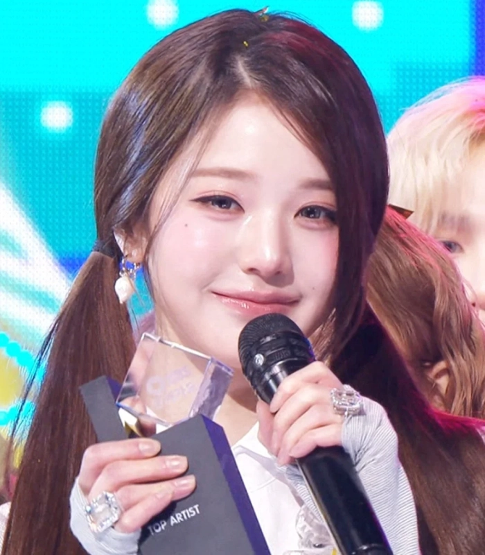 K-POPアイドルと思われる女性が、ステージ上で「TOP ARTIST」と書かれたトロフィーとマイクを持ち、笑顔を見せている。ツインテールの髪型で、アクセサリーを身につけており、授賞式での喜びが伝わる瞬間を捉えた一枚です。