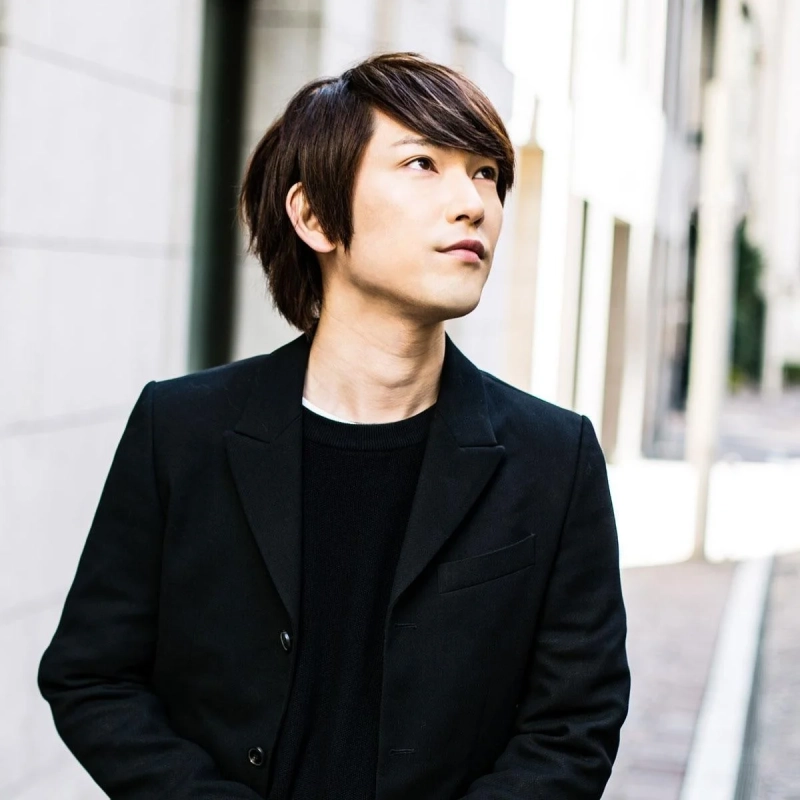 黒いジャケットとセーターを着用したアジア系の若い男性が、屋外のストリートで上を見上げているポートレート写真です。