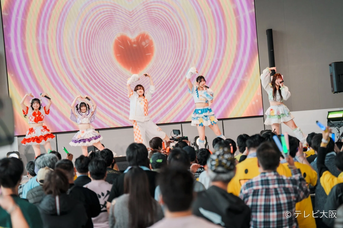 ハート柄の大型スクリーンを背景にパフォーマンスするアイドルグループ