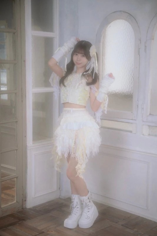 白いレースとフェザーの衣装を身につけた女性アイドルが、明るいスタジオで微笑んでいる全身ポートレート。