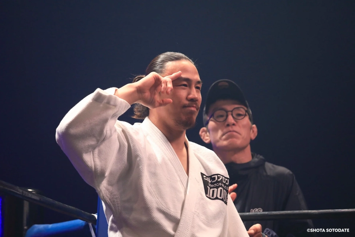 白い柔術着を着た男性がリング上でポーズをとっており、その後ろには黒い服を着た男性が立っている。