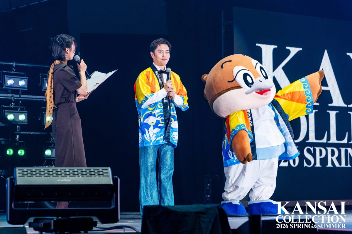 女性司会者、和装の男性プレゼンター、そしてカラフルな衣装を着たマスコットキャラクターが並んでいます