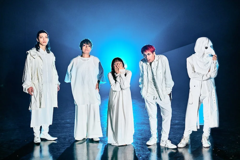 青い光が当たる暗い背景の中、白を基調とした個性的な衣装を身につけた5人が一列に並んで立っています