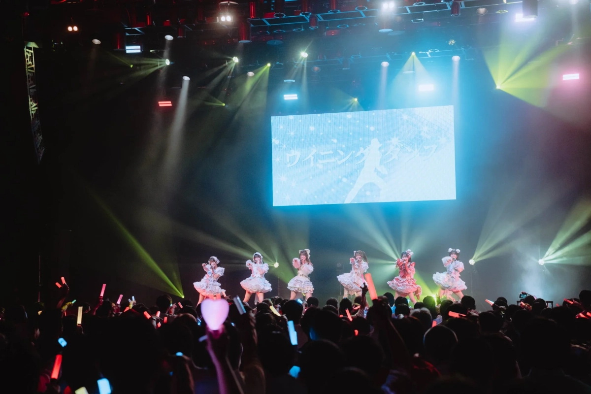 複数のアイドルが華やかなステージでパフォーマンスしており、大勢の観客がペンライトを振って盛り上がっているライブ会場の様子。背景のスクリーンには「ウィニング」の文字が見える。