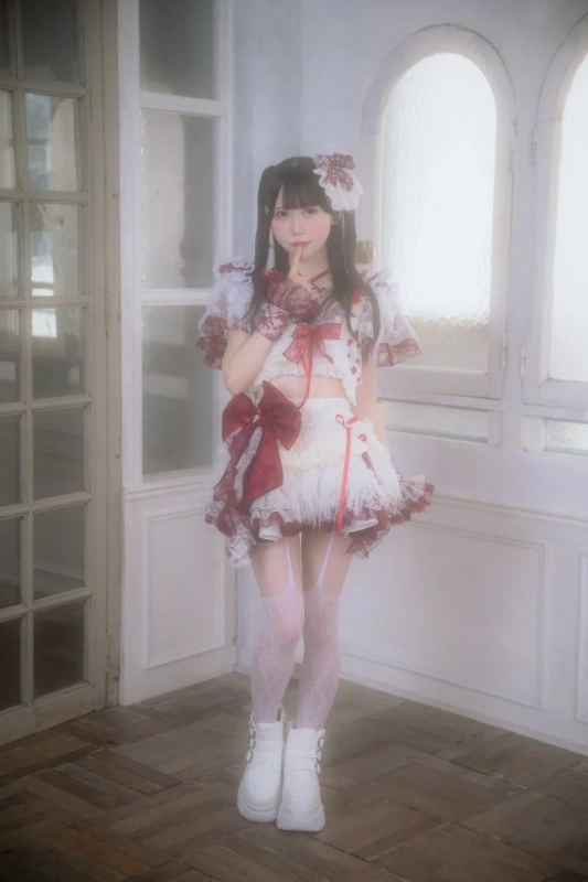 白と赤のフリルが特徴的なかわいらしい衣装をまとった女性が、室内でポーズをとっています。