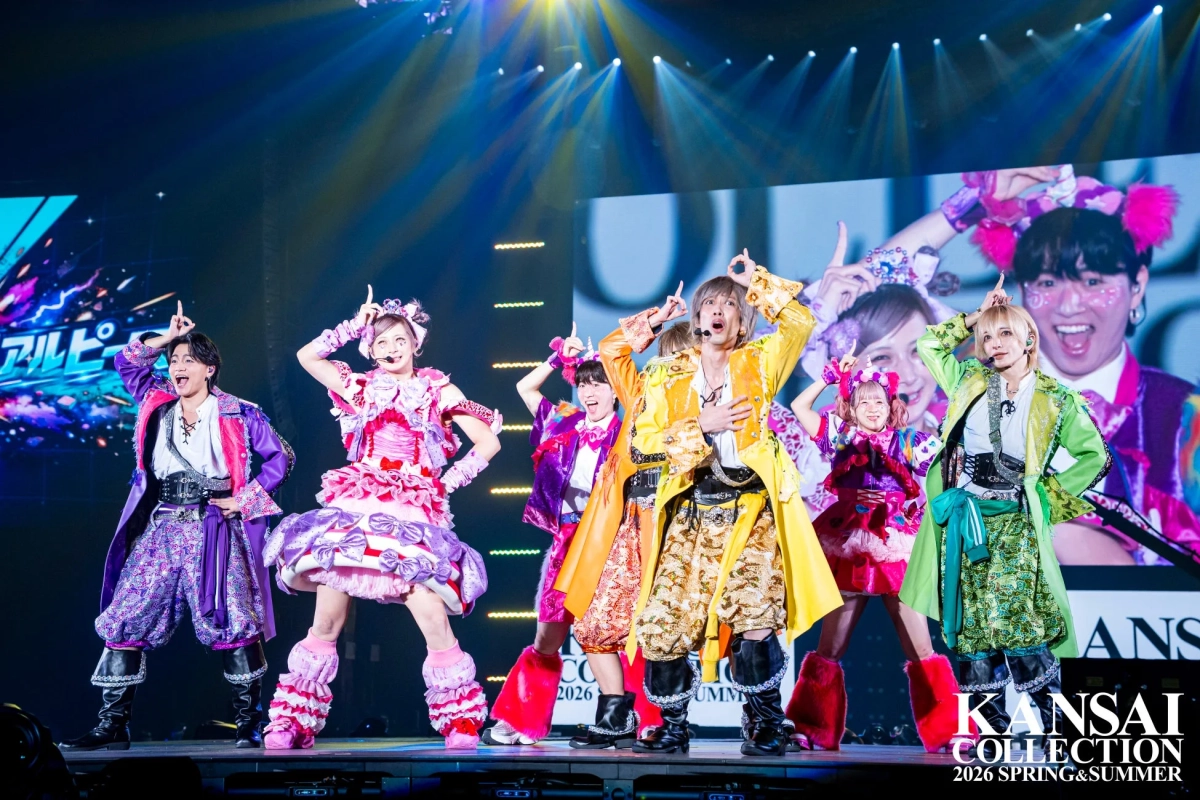 カラフルで個性的な衣装を身につけたパフォーマーたちが、明るく照らされたステージ上で活気あふれるポーズをとっています