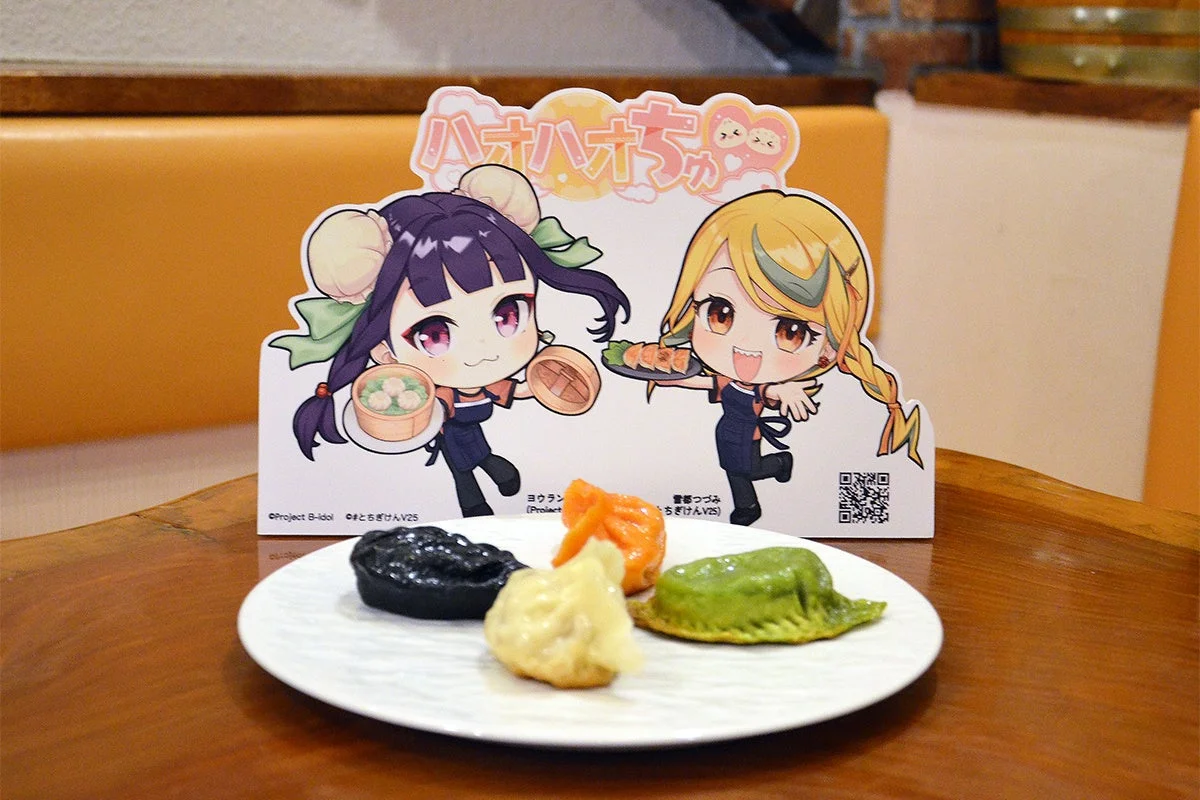 アニメキャラクターのパネルが置かれたテーブルに、黒、オレンジ、白、緑色のカラフルな餃子や点心が盛り付けられた皿がある様子