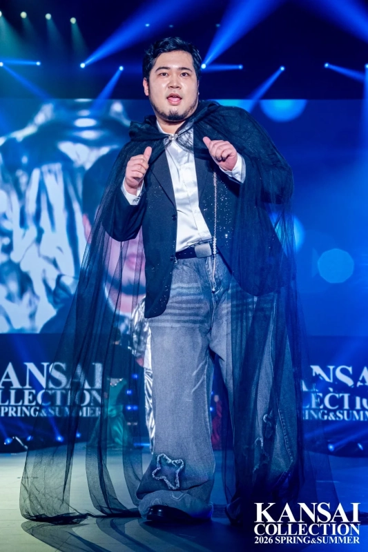 黒いケープとジャケット、デニムを着用した男性がポーズをとっています