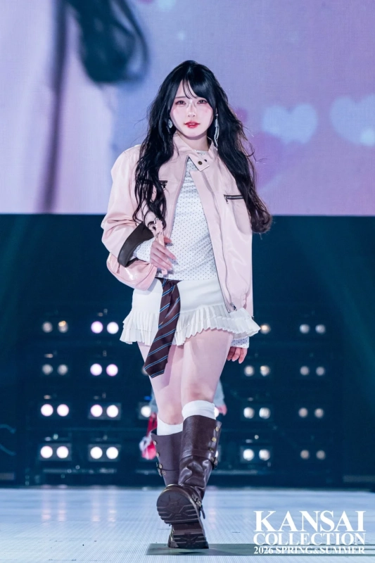 ピンクのライダースジャケットと白いミニスカート、茶色のロングブーツを着用した若い女性モデルが、ランウェイを歩いている様子