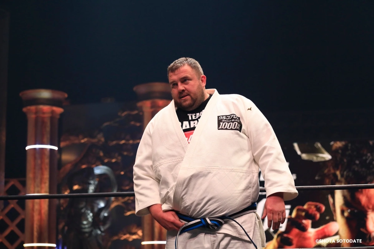 白い道着と青帯を着用した男性が格闘技のリング内に立っている様子。