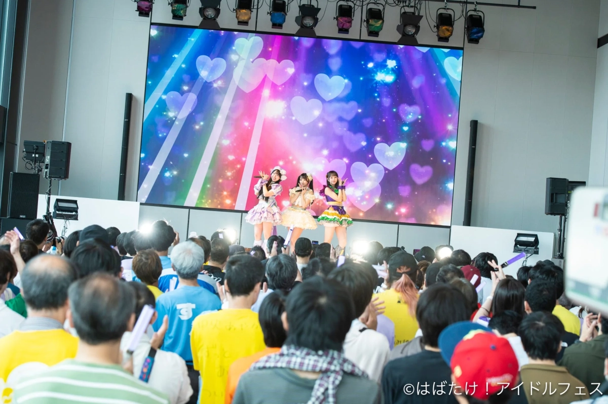 カラフルな衣装を着た3人のアイドルが、ハート模様の大型LEDスクリーンを背景にステージでパフォーマンスしており、多くの観客がそれを見守るライブイベントの様子です。