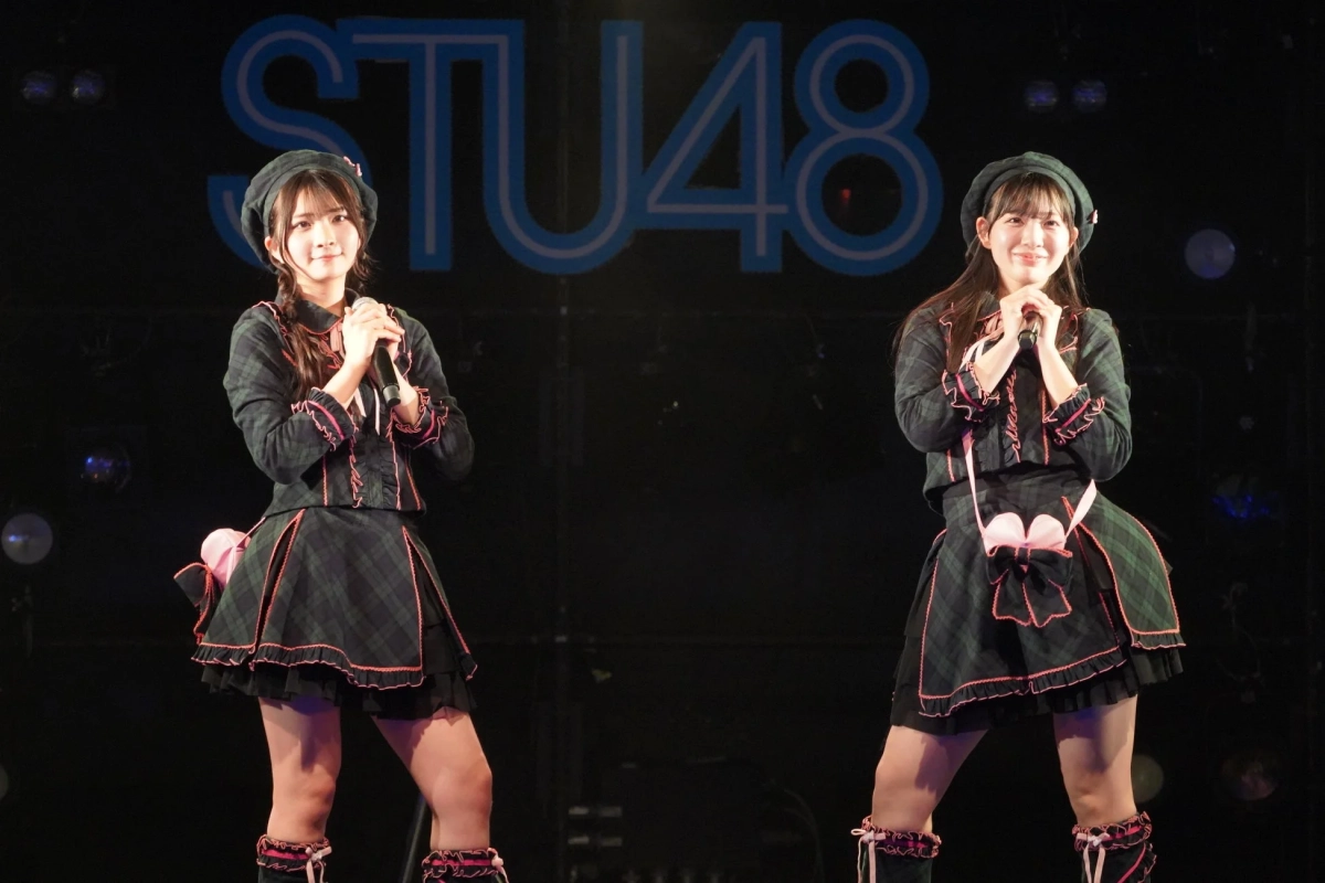 STU48のメンバーと思われる2人の女性アイドルが、チェック柄の衣装とベレー帽を着用し、マイクを持ってステージ上で笑顔でポーズをとっています