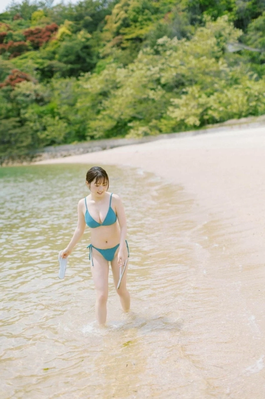 青いビキニを着た若い女性が、白いサンダルを手に持ち、日差しの降り注ぐビーチの浅瀬を歩いています