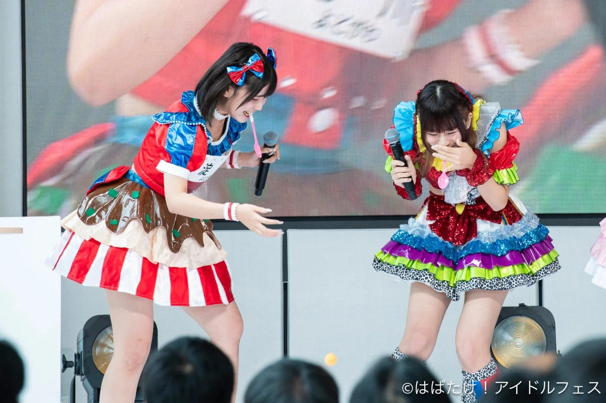カラフルな衣装をまとった2人のアイドルがステージ上で楽しそうにしている様子。一人はマイクを持ち、もう一人は笑いながら口元を隠している。ピンポン玉が床に落ちており、ゲーム中の賑やかな一幕を捉えた一枚。