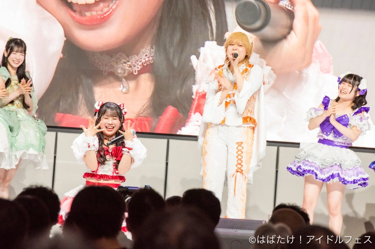 カラフルな衣装を身につけたアイドルグループがステージでパフォーマンスをしている様子。メンバーは笑顔で歌ったり踊ったりしており、観客も楽しんでいる活気あるライブイベントの瞬間を捉えている。