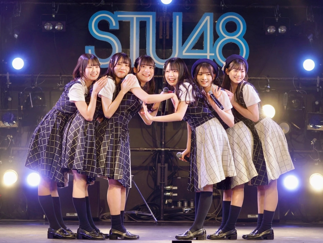 ライブステージで、チェック柄の衣装を着たSTU48のメンバー6人が笑顔でポーズをとっています