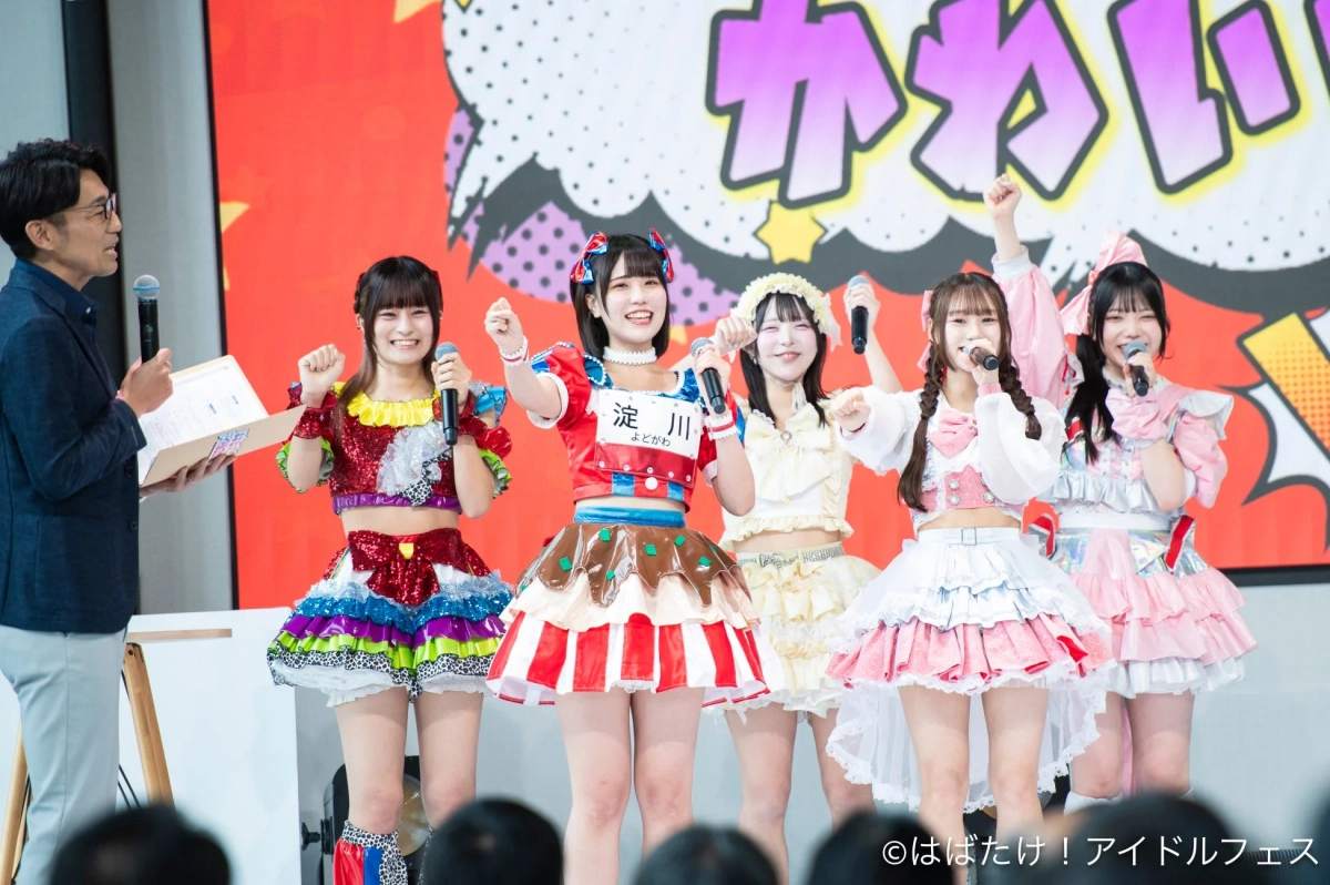 カラフルな衣装を身につけた5人の女性アイドルグループがステージでパフォーマンスしている様子。司会者もおり、「かわいい」と書かれた背景の前で笑顔を見せ、マイクを持って歌っている。アイドルフェスの熱気が伝わる一枚。