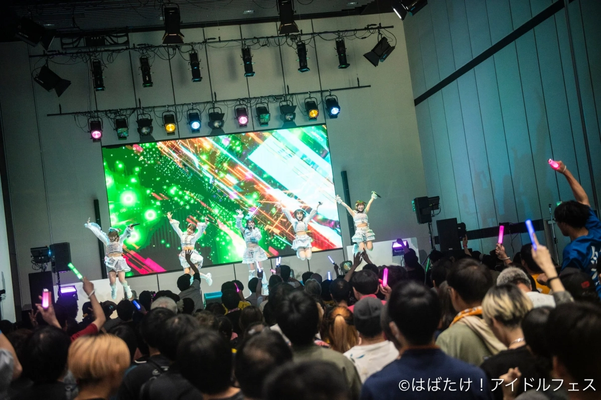 アイドルグループがステージでパフォーマンスしており、観客がサイリウムを振って応援しているライブ会場の様子です。メンバーがジャンプしている躍動感のある瞬間を捉えています。