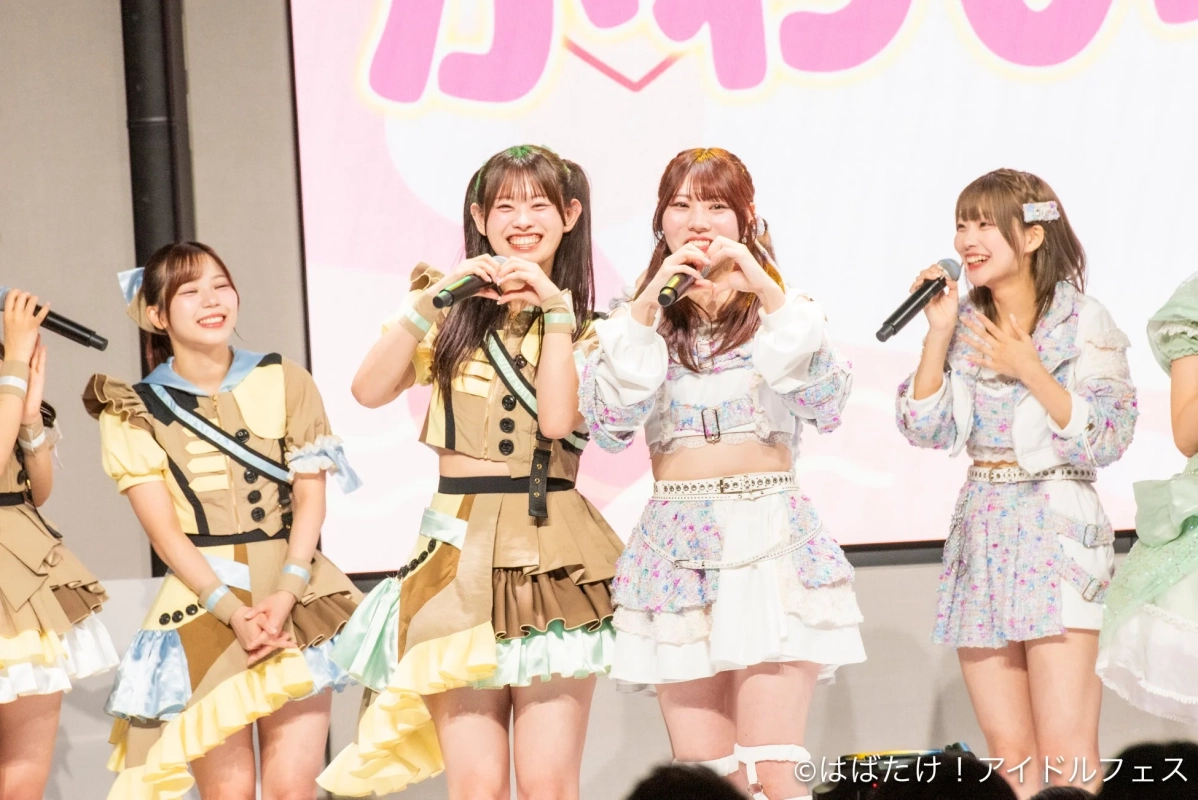 複数の女性アイドルがステージ上で笑顔でパフォーマンスしている様子。マイクを手に歌い、ハートのポーズをとるメンバーもいる。華やかな衣装を身につけ、楽しげな雰囲気のライブイベントの一幕である。
