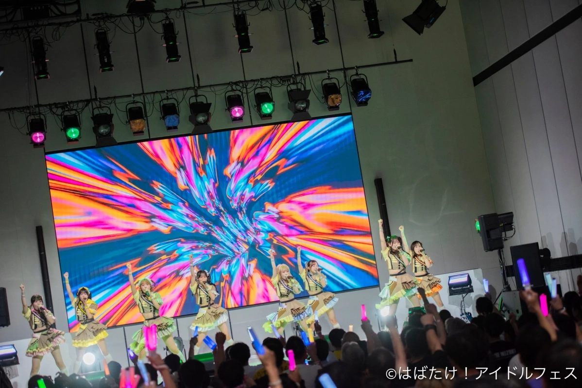 カラフルなLEDスクリーンを背景に、女性アイドルグループがステージでエネルギッシュなパフォーマンスを披露しています。観客はサイリウムを振って応援しており、会場は熱気に包まれています。
