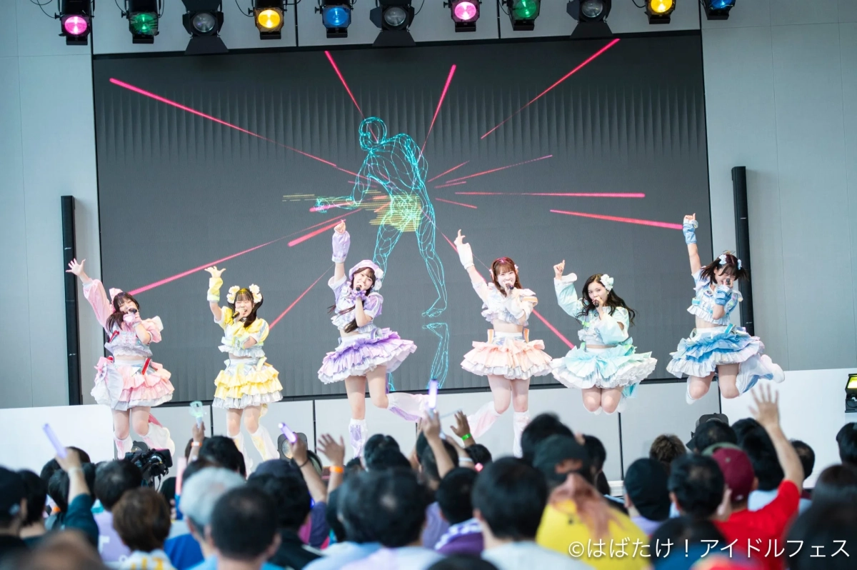 カラフルなフリルの衣装を着た6人の女性アイドルグループが、ステージ上でジャンプしながらパフォーマンスを披露しています。背景の大型スクリーンにはデジタルな光の演出が映し出され、多くの観客がペンライトを振って盛り上がっています。