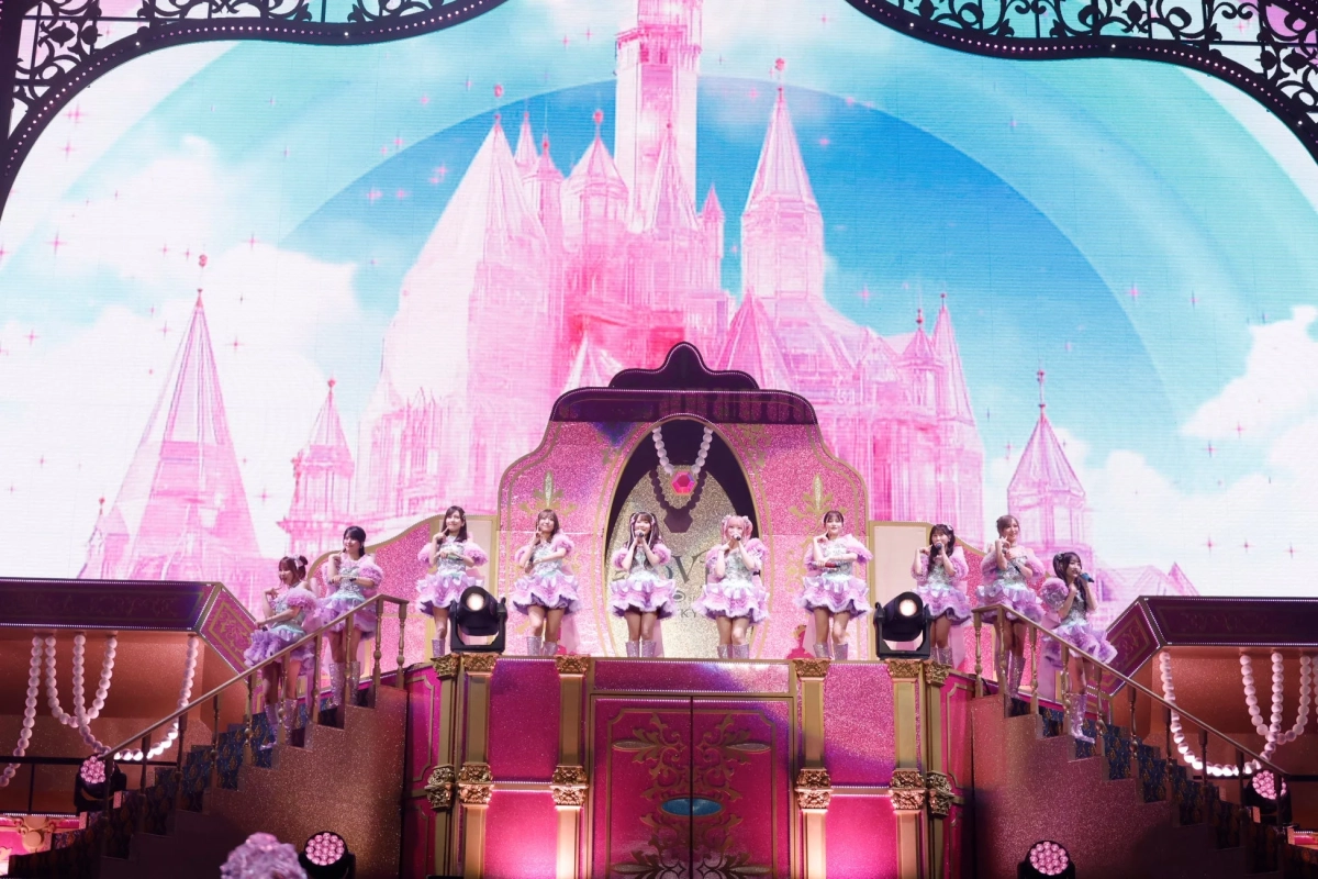 ピンクの城が背景のファンタジックなステージで、複数の女性アイドルが可愛らしい衣装を着てパフォーマンスを行っている様子
