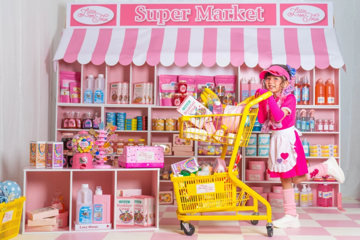 ピンクと白の可愛らしいスーパーマーケットで、ピンクの衣装の少女が黄色いショッピングカートを押している