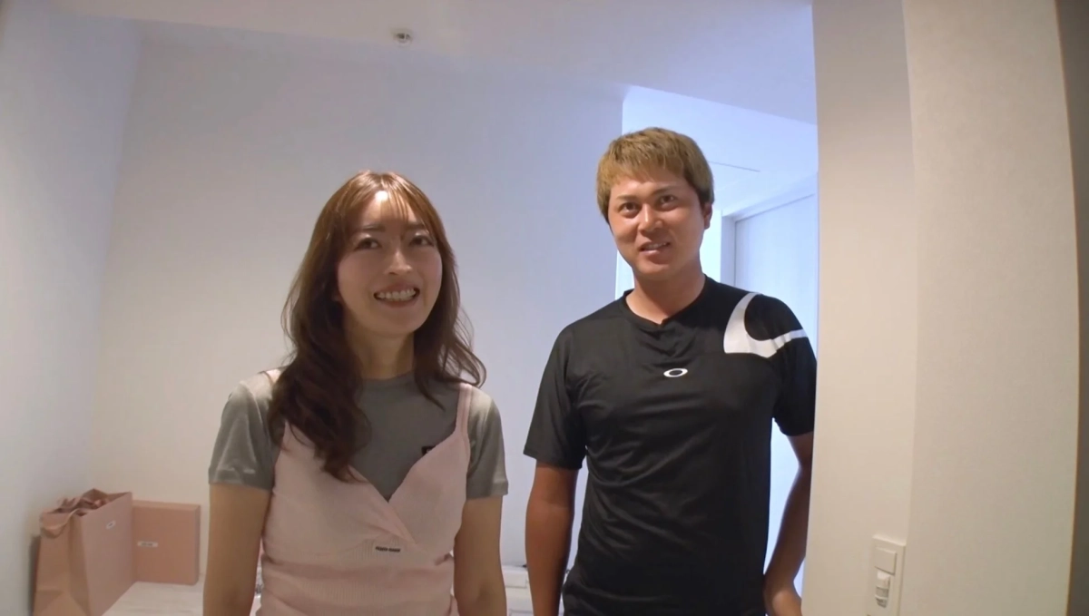 白い部屋で男女二人が笑顔でカメラを見ている。女性はピンクのトップス、男性は黒いTシャツを着用しており、カジュアルな雰囲気。背景には茶色の紙袋が見える。