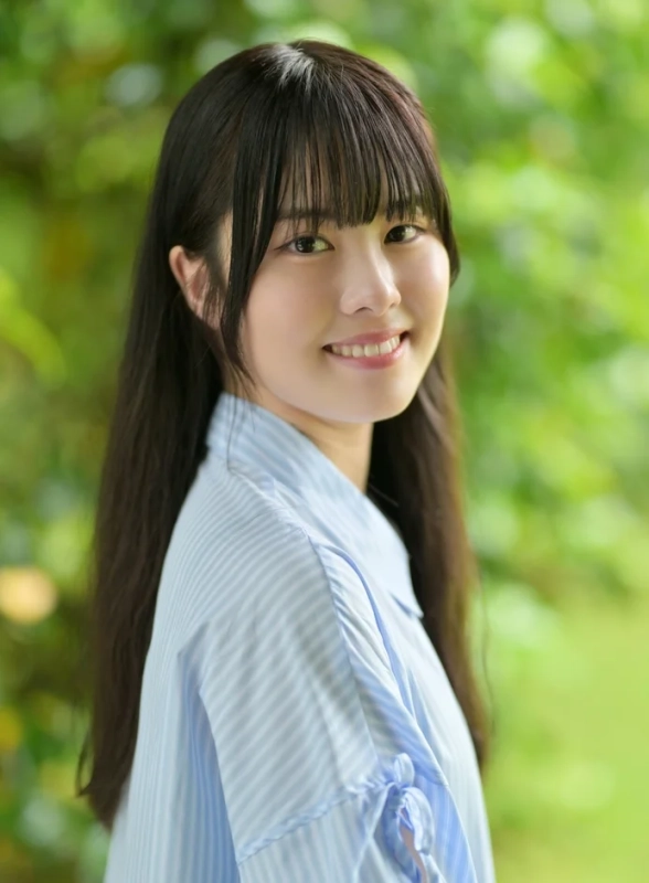 緑豊かな背景を背に、水色のストライプシャツを着た若い女性がカメラに向かって優しく微笑んでいます
