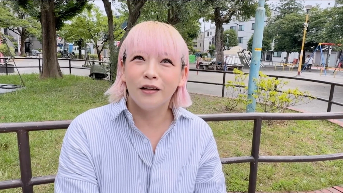ピンクと金髪のツートーンカラーの髪をした女性が、屋外の公園でストライプのシャツを着て、何かを見上げるような表情をしている瞬間を捉えた画像です。