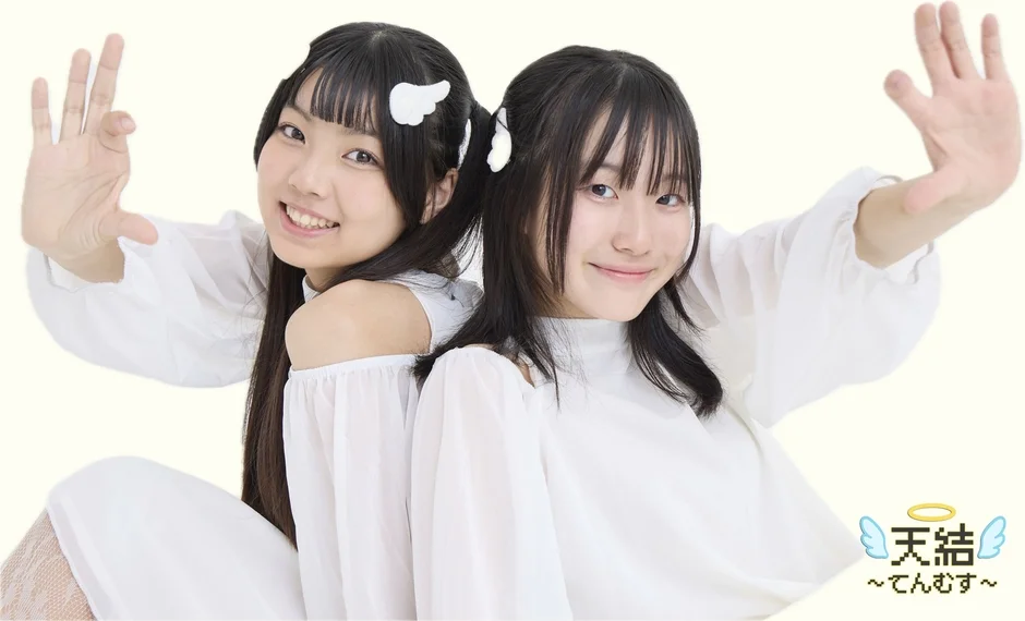 白い衣装を着た2人の若い女性が背中合わせで笑顔でポーズをとっています