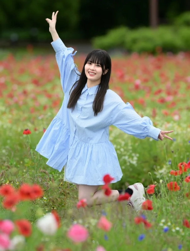 赤いポピーが咲く花畑で、青いワンピースを着た若い女性が笑顔で両腕を広げ、楽しそうにポーズをとっています