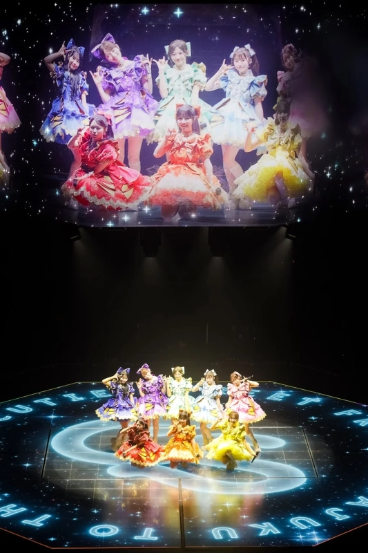 カラフルでフリルの多い衣装をまとったアイドルグループが、光る文字と模様が描かれたステージでパフォーマンスをしている様子です。背景の大きなスクリーンにもメンバーの姿が映し出されており、華やかで活気あるライブの雰囲気が伝わってきます。