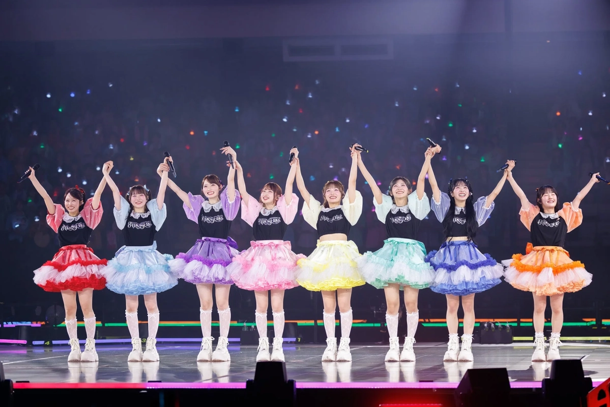 8人組の女性アイドルグループがステージ上で手を繋ぎ、腕を上げて笑顔で観客に手を振っている様子。カラフルなフリルスカートの衣装を着用しており、ライブ会場の熱気が伝わってくる一枚です。