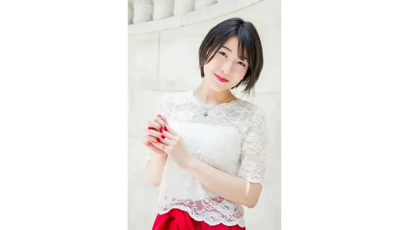 ショートヘアの若い女性が白いレースのトップスと赤いスカートを着用し、優しく微笑んでいます。明るい壁を背景にポーズをとっており、清楚で可愛らしい印象のポートレートです。