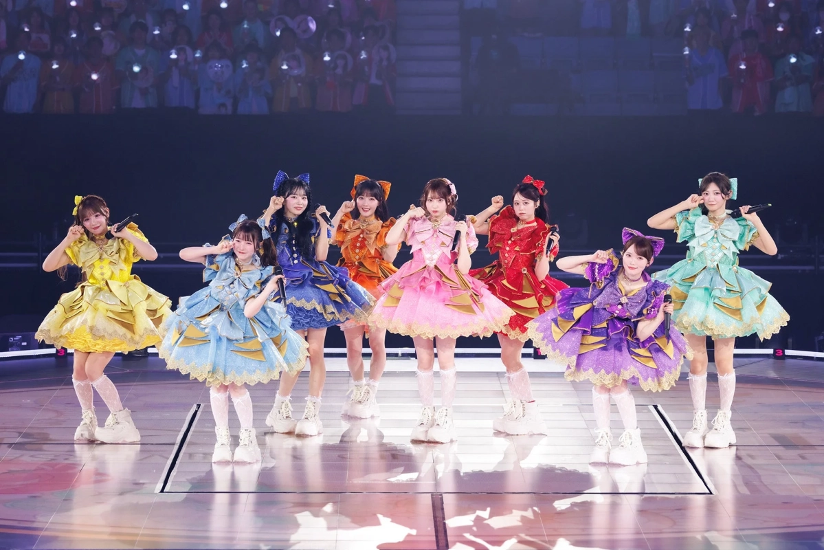 華やかなフリルのドレスをまとった8人の女性アイドルがステージでパフォーマンスしている様子。背景にはペンライトを持つ大勢の観客が見える。