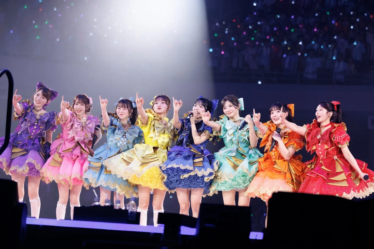 カラフルなアイドル衣装をまとった8人の女性グループが、ステージで笑顔でパフォーマンスしているライブの様子。観客のペンライトが輝く中、メンバーは歌い、指をさすポーズで会場を盛り上げている。