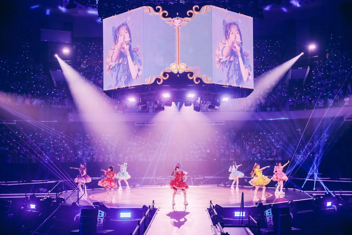 華やかなステージで複数の女性アイドルがパフォーマンスを行っているライブコンサートの様子です。会場には多くの観客がペンライトを振っており、ステージ上にはメンバーの姿を映し出す巨大スクリーンと、鮮やかな照明やレーザーが演出されています。