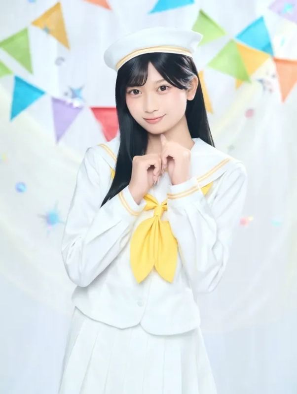 白いセーラー服と帽子を着用した女性が、黄色いリボンを胸元につけ、穏やかな笑顔でポーズをとっています。背景にはカラフルな三角旗が飾られており、明るく華やかな雰囲気です。